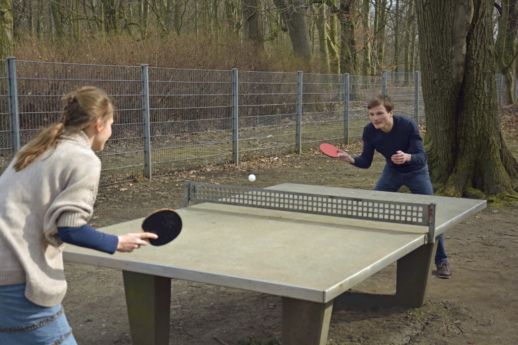 An einer Tischtennisplatte im Park spielen eine Frau und ein Mann.