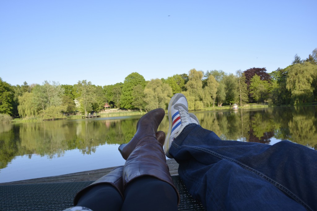 Die Beine einer männlichen und einer weiblichen Person auf einem Steg vor einem See. 