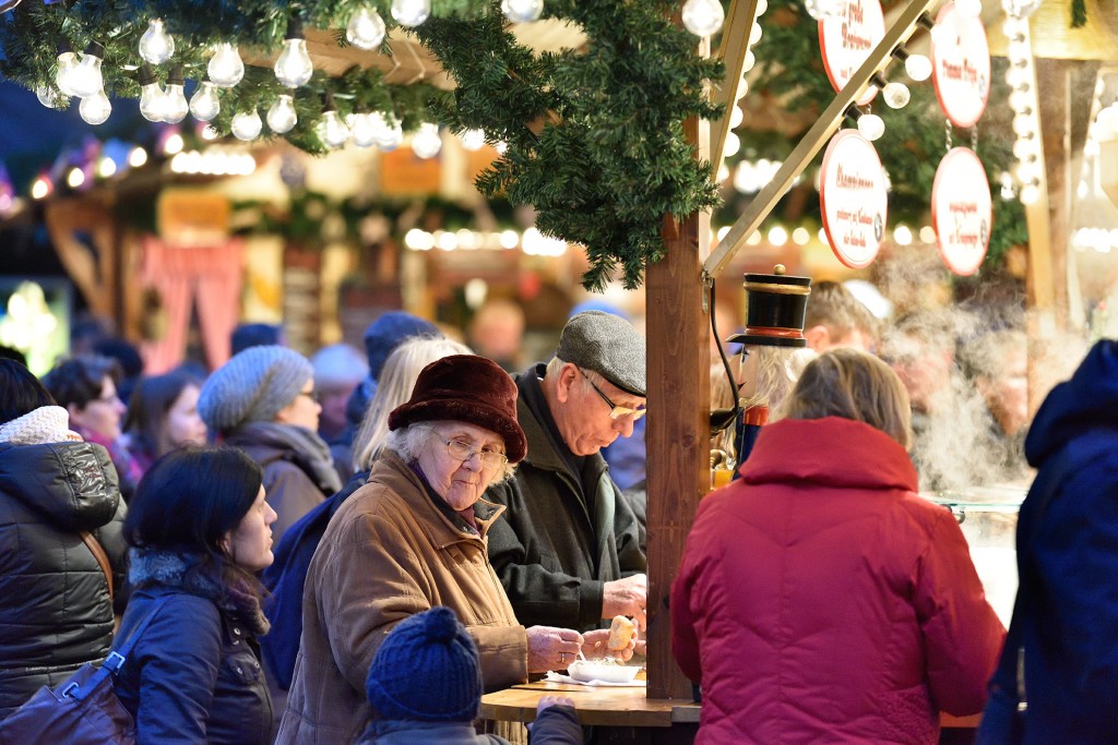 An einer Weihnachtsmarktbude steht ein älteres Ehepaar und isst, die Dame schaut in die Kamera.