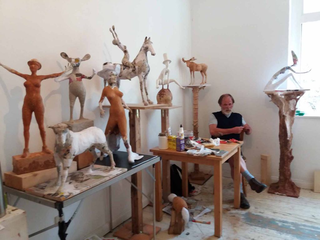 Ein Künstler sitzt auf einem Stuhl inmitten seiner Skulpturen. Die Skulpturen sidn verschiedene Tiere und menschliche Körper.