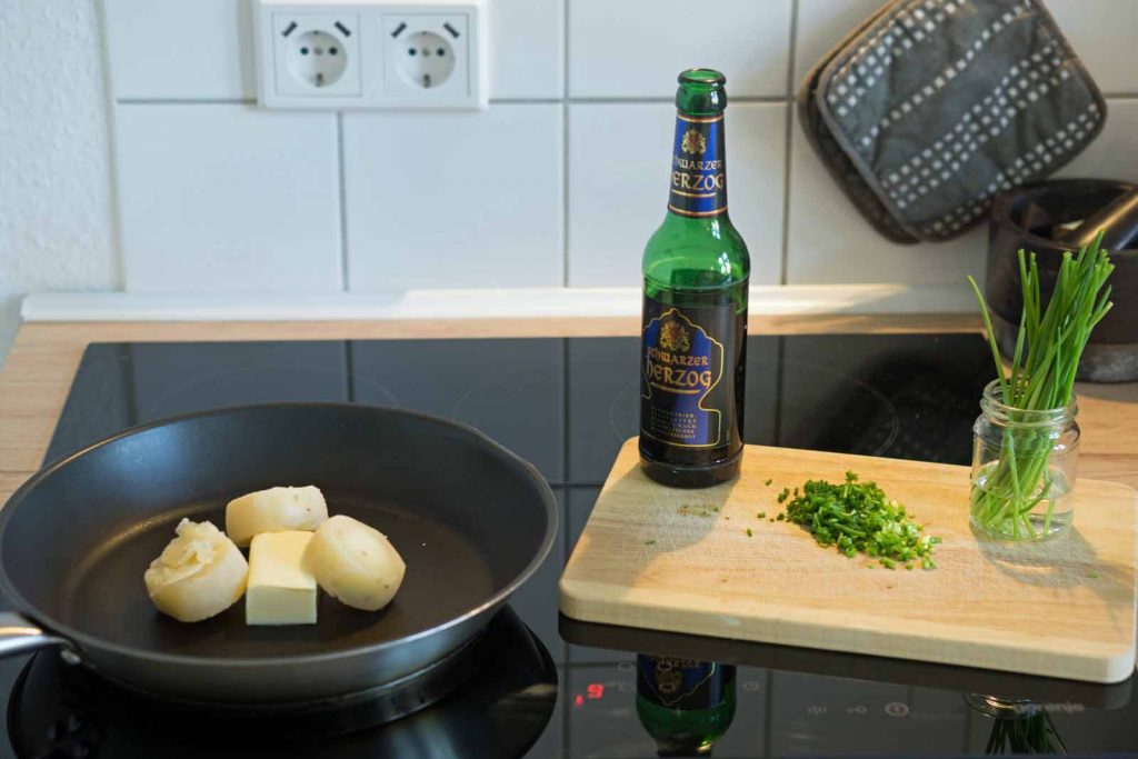 Auf einem Herd steht eine Pfanne mit Harzer Roller und Butter, daneben eine Flasche Schwarzer Herzog und kleine Schnittlauchröllchen.