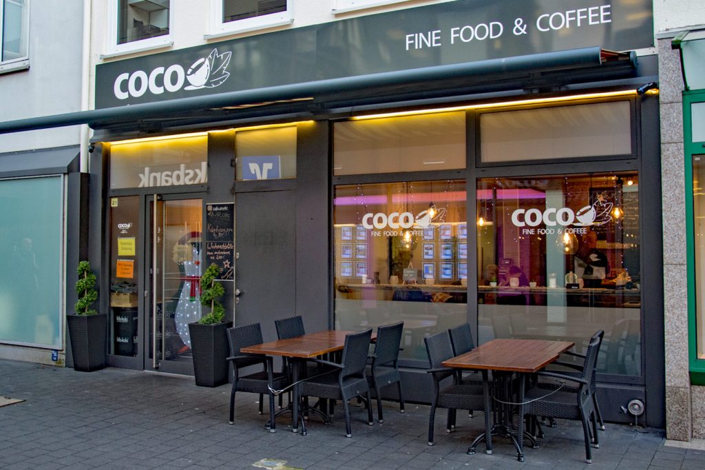 Die Außenfront des Café Coco. Vor den Fenstern stehen zwei Tische mit Stühlen.