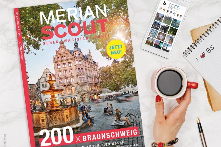 Das Heft MERIAN Scout Braunschweig liegt auf einem Tisch, daneben eine Hand, die einen Kaffee hält, ein Handy und ein Notizblock.