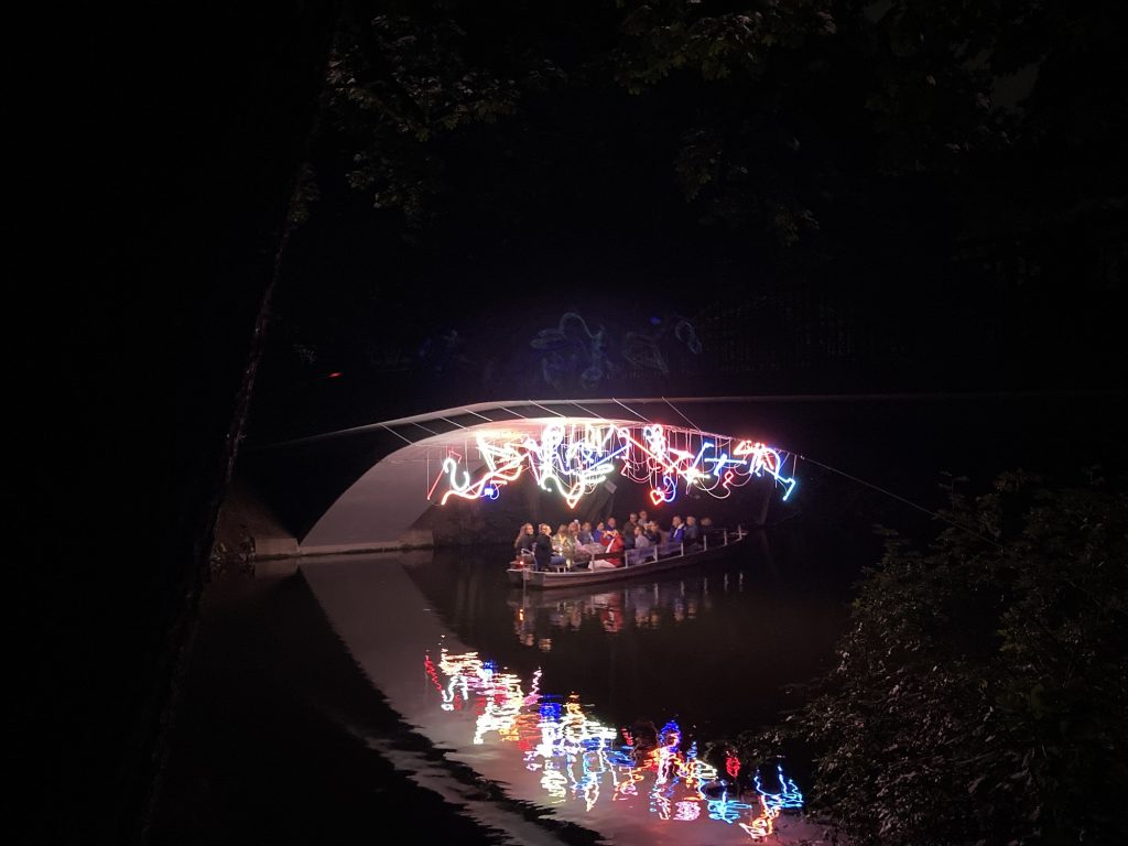 Bunte Lichter unter einer Brücke. Darunter ein Kahn mit Menschen. 