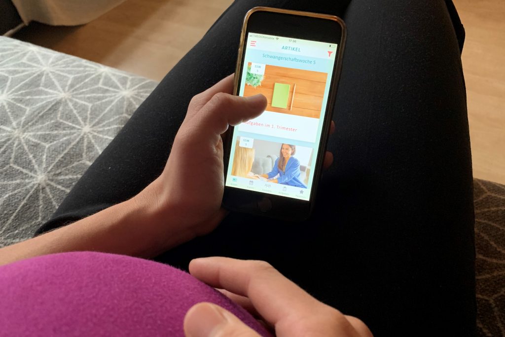 Eine schwangere Frau sitzt auf einem Sofa, in ihrer Hand ein Handy mit der Babybauch App