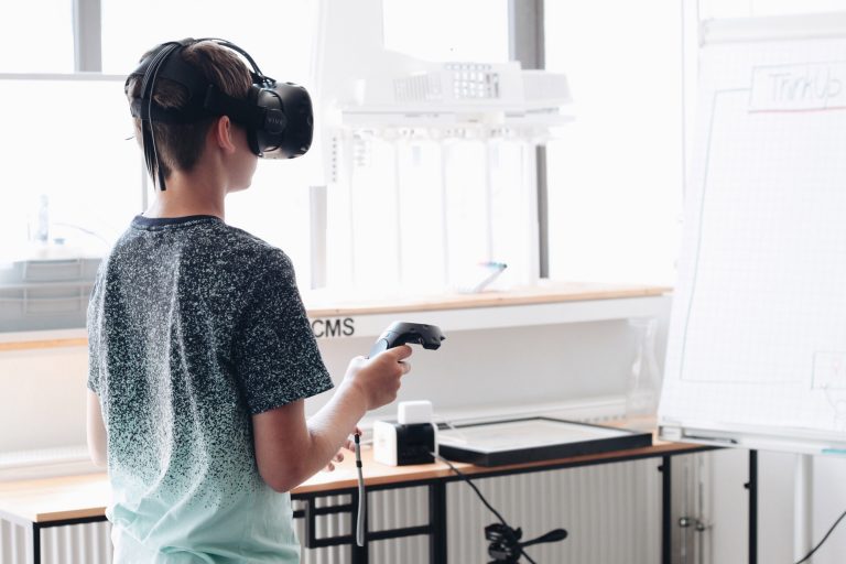 Ein Junge im T-Shirt steht in einem Klassenraum. Er hat eine VR-Brille auf dem Kopf und hält einen Controller in der Hand.