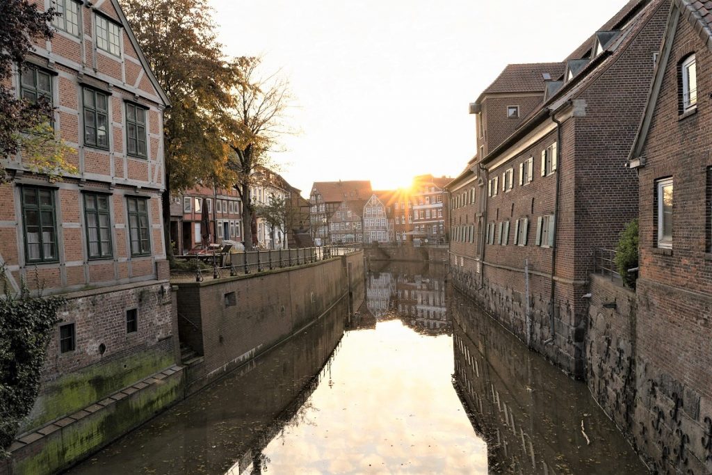 An einem Kanal stehen Wohnhäuser. Über den Häusern bricht die Sonne und taucht die Stadt in ein goldenes Licht.