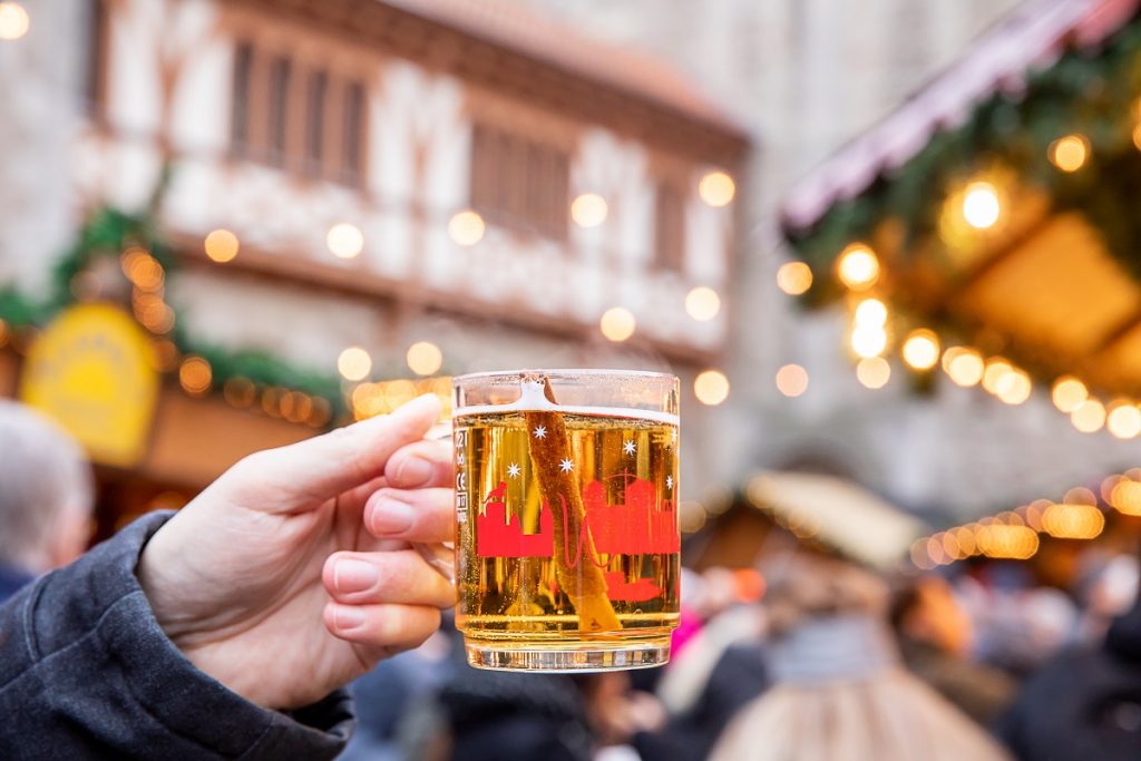 Ein Glas gefüllt mit Apfelglühwein und Zimtstange im Vordergrund, im Hintergrund verschommen der Übergang von Dom zur Burg und Weihnachtsmarktstände.