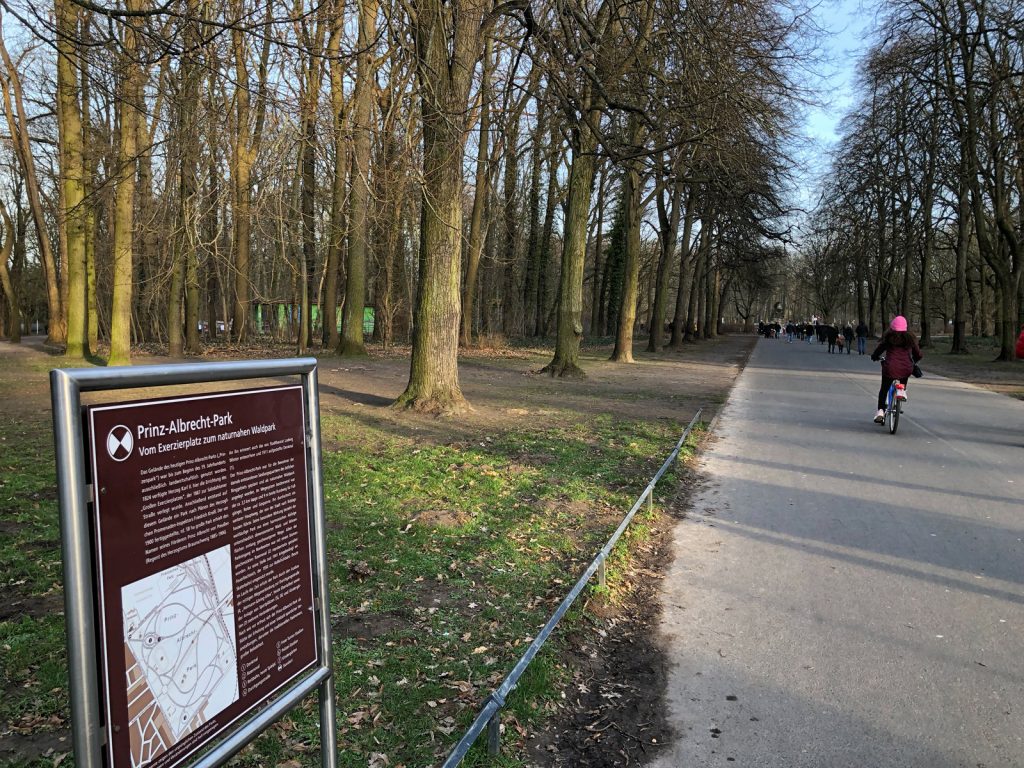 Rechts ein geteerter Weg in einen Park hinein, links vorne im Bild eine Tafel mit Informationen zum Prinz-Albrecht-Park sowie einem Lageplan.