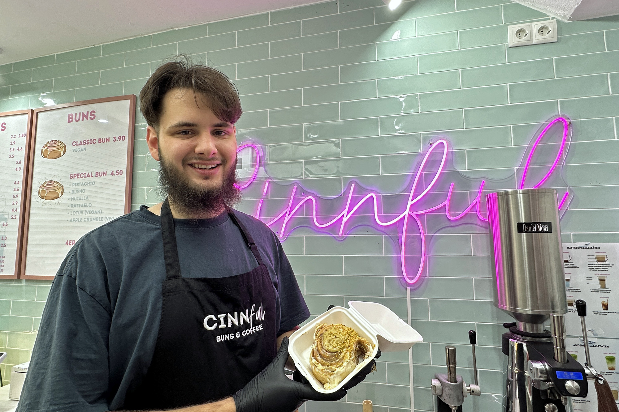 Ein Mann hält vor dem Ladenschild von Cinnful eine Zimtschnecke lächelnd in die Kamera.