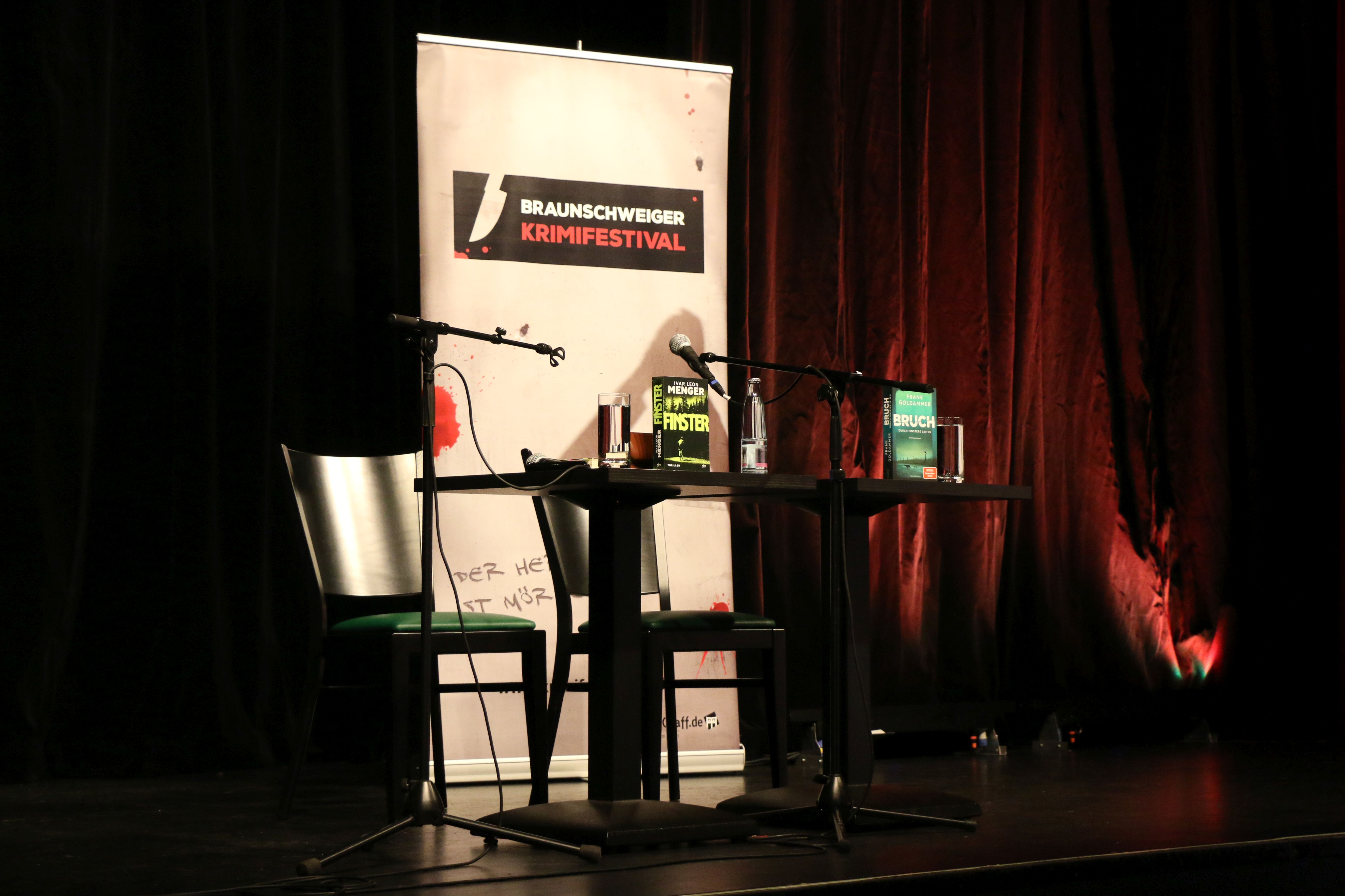 Tisch mit zwei Stühlen und Mikrofonen vor einer Leinwand auf der "Braunschweiger Krimifestival" steht.